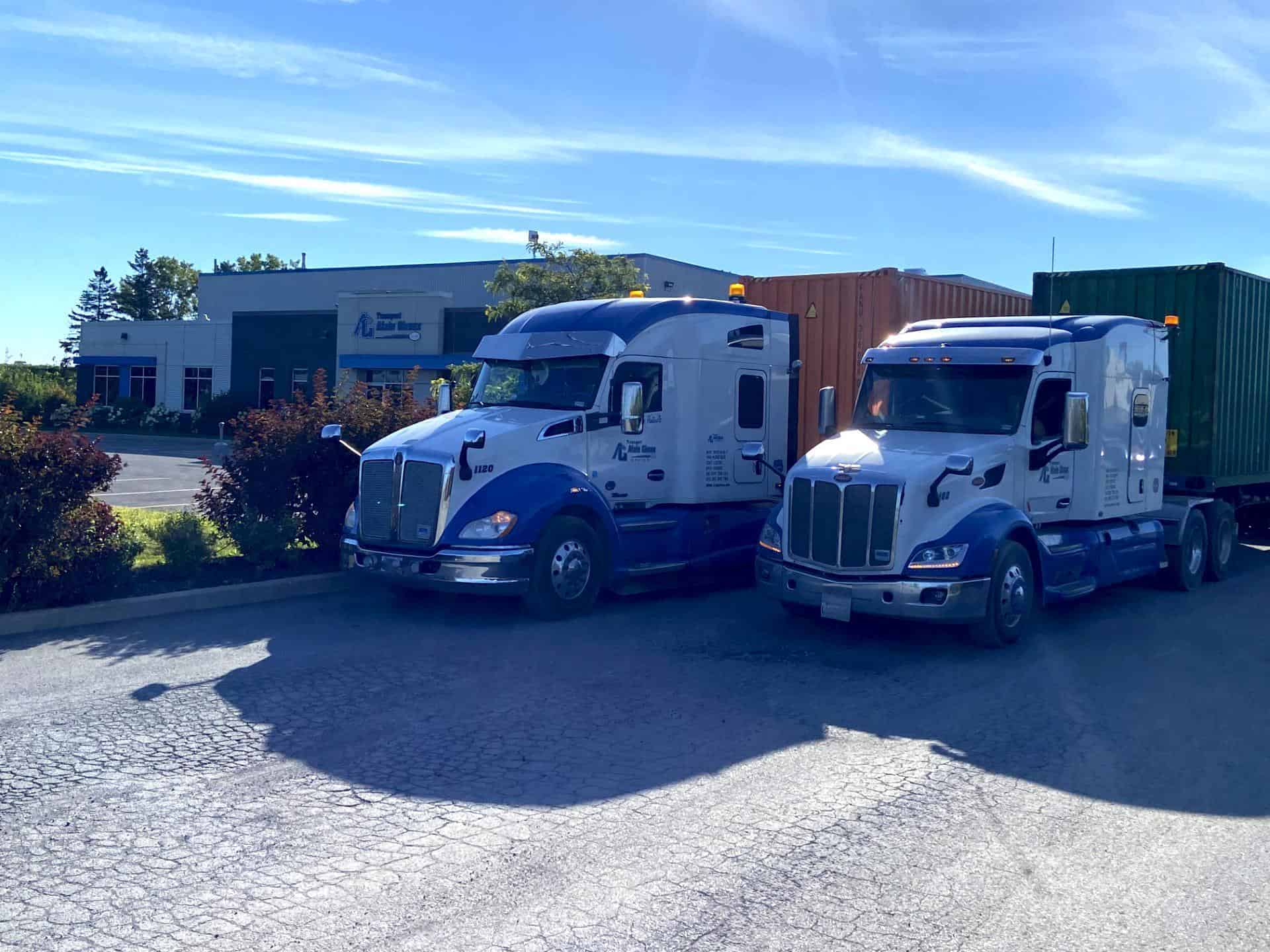 Deux semi-remorques bleus et blancs sont garés côte à côte sur un terrain près d'un bâtiment commercial, chacun transportant un conteneur d'expédition.