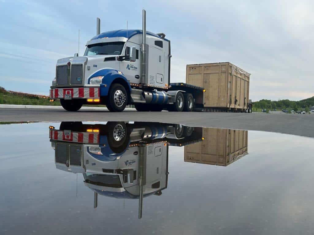 A blue and white semi-truck hauling a large wooden crate is reflected in a puddle on an overcast day, parked on an asphalt surface.