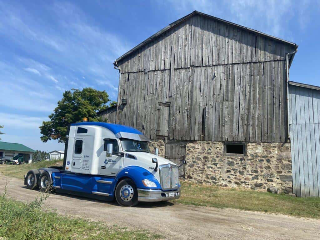A blue and white semi-truck is parked on a dirt road beside a large, weathered wooden barn under a partly cloudy sky.