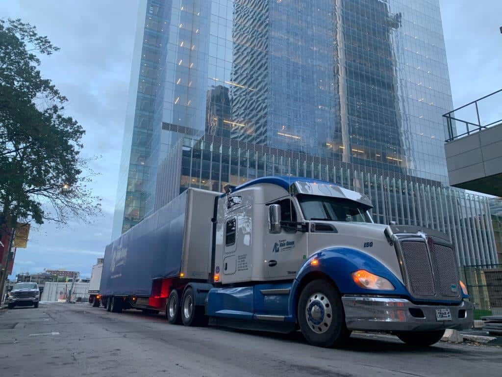 A large blue and white semi-truck is parked on a city street in front of a tall glass office building.
