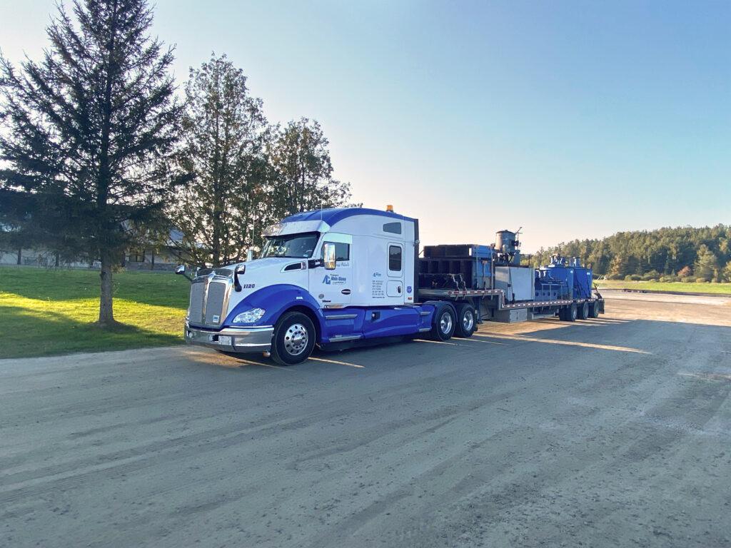 A white and blue semi-truck with a flatbed trailer carrying industrial equipment is parked on a dirt lot near grass and trees under a clear sky.