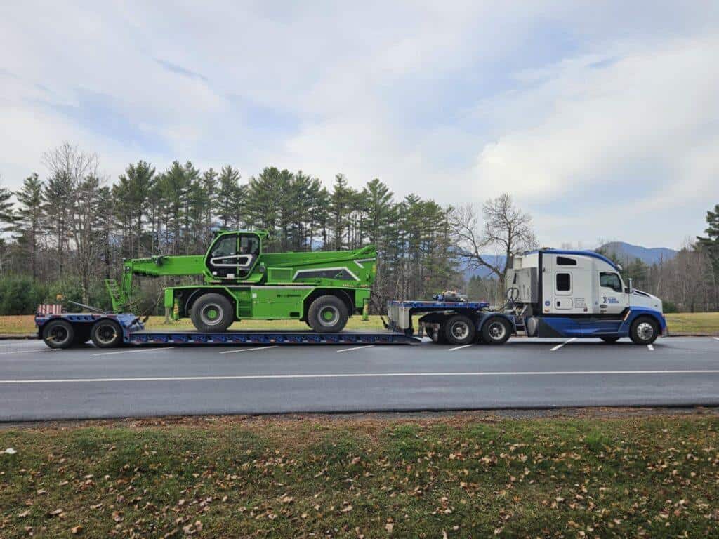 A white semi-truck transports a bright green construction vehicle on a flatbed trailer along a road lined with trees.